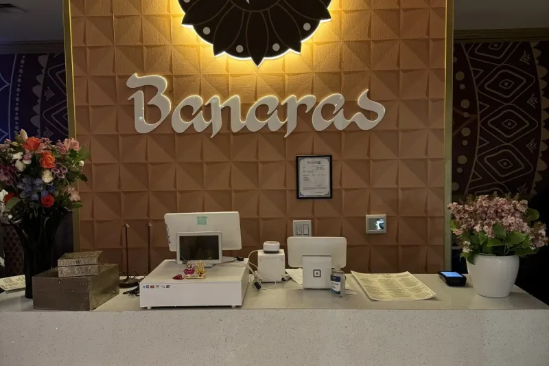The host counter at Banaras in Bellevue, with the restaurant's illuminated flower-logo sign and 'Banaras' lettering on a copper-tiled wall above a stone counter with flower arrangements