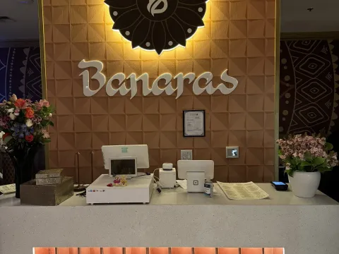 The host counter at Banaras in Bellevue, with the restaurant's illuminated flower-logo sign and 'Banaras' lettering on a copper-tiled wall above a stone counter with flower arrangements