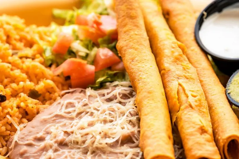 A plate of crispy rolled flautas with Mexican rice, refried beans topped with shredded cheese, salad and sides of dressing and guacamole at Delicious Mexican Eatery in El Paso