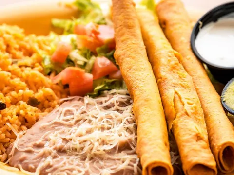 A plate of crispy rolled flautas with Mexican rice, refried beans topped with shredded cheese, salad and sides of dressing and guacamole at Delicious Mexican Eatery in El Paso