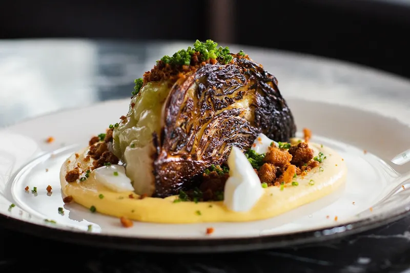 Charred wedge of cabbage over a swoosh of purée, topped with herbs and crispy crumbs, plated on a white dish at Sắp Sửa in Denver