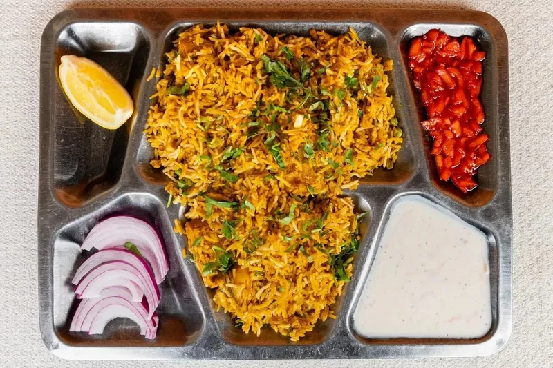 A biryani thali on a steel compartment tray at Punjabi Dhaba in Cambridge, with saffron-hued rice, sliced red onion, chopped tomato and raita