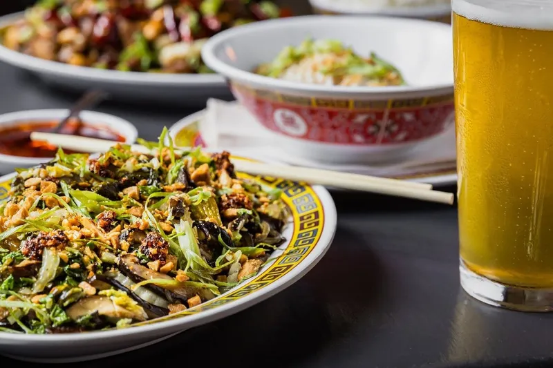A spread of Sichuan dishes at Kwei Fei in Charleston — stir-fried greens and mushrooms topped with chili crisp and peanuts on a classic Chinese-pattern plate, a bowl of steamed rice, and a glass of beer
