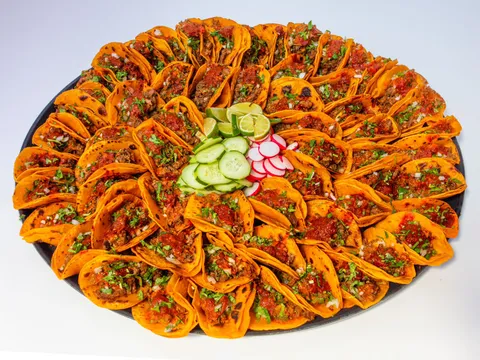 A large round party platter of Birria-Landia beef birria tacos on orange corn tortillas garnished with cilantro, onion and red salsa, ringed around sliced cucumber, radish and lime